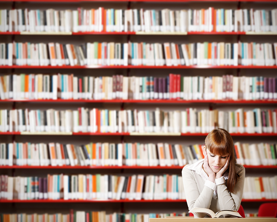 Una estudiante lee un libro frente a una estantería de grandes dimensiones repleta de tomos de diferentes géneros